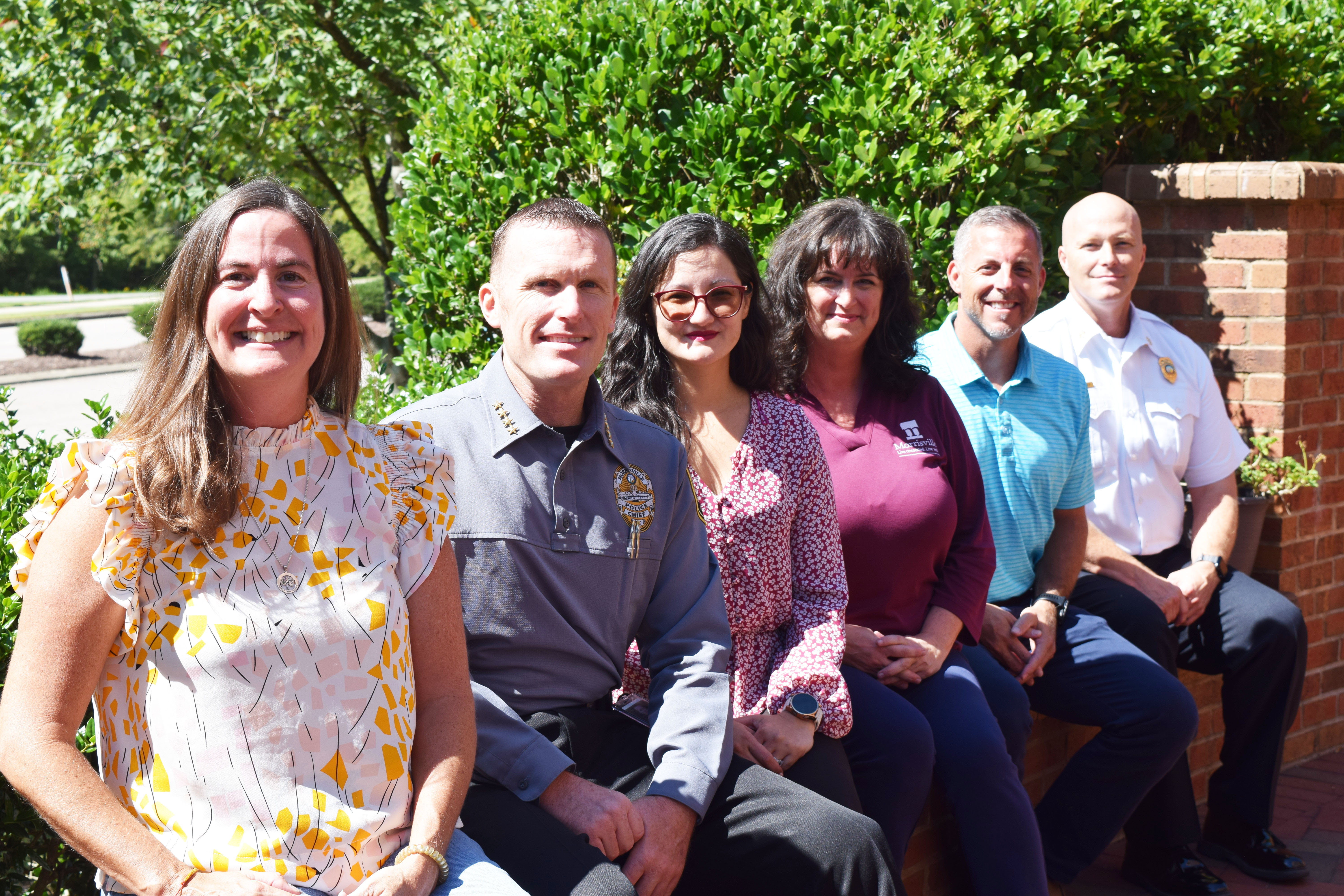 A group of six adults seated side by side on a low brick wall smiling at the camera with the sun shining on them