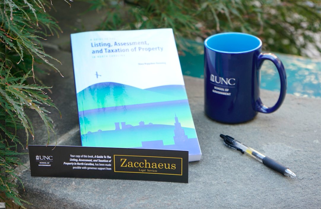 A textbook titled "Listing, Assessment, and Taxation of Property in North Carolina" and a bookmark branded with the Zacchaeus Legal Services logo sit next to greenery, a black ink pen, and a UNC School of Government mug.