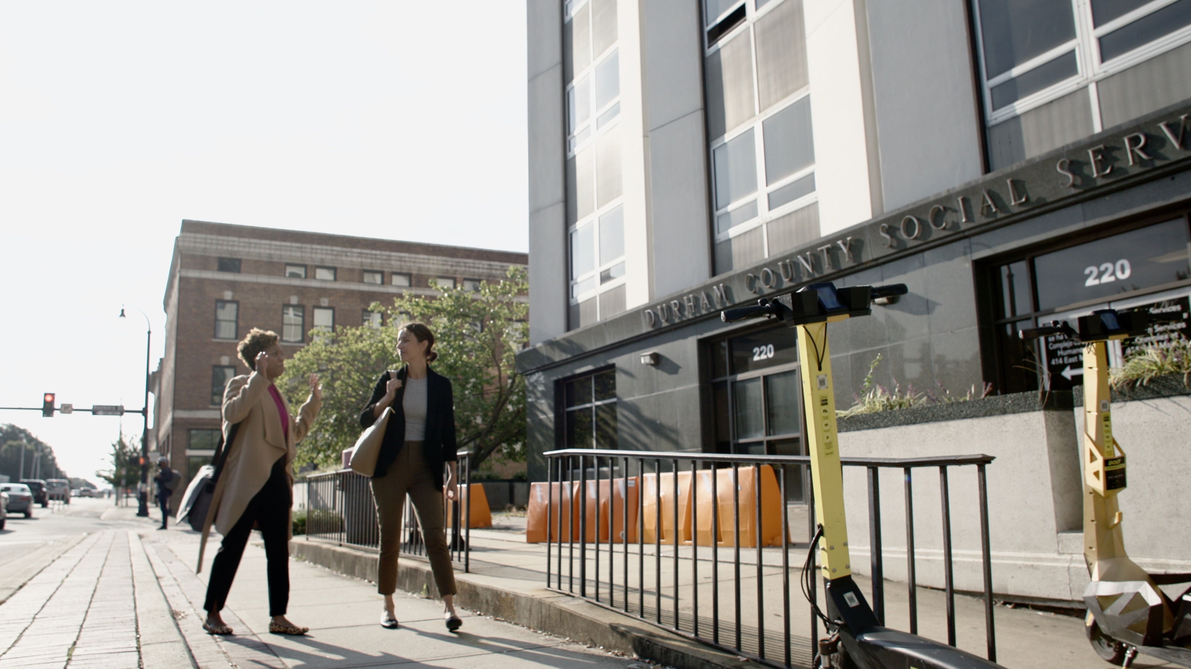 Two women walk while talking on a sidewalk next to the Durham County Social Services building.