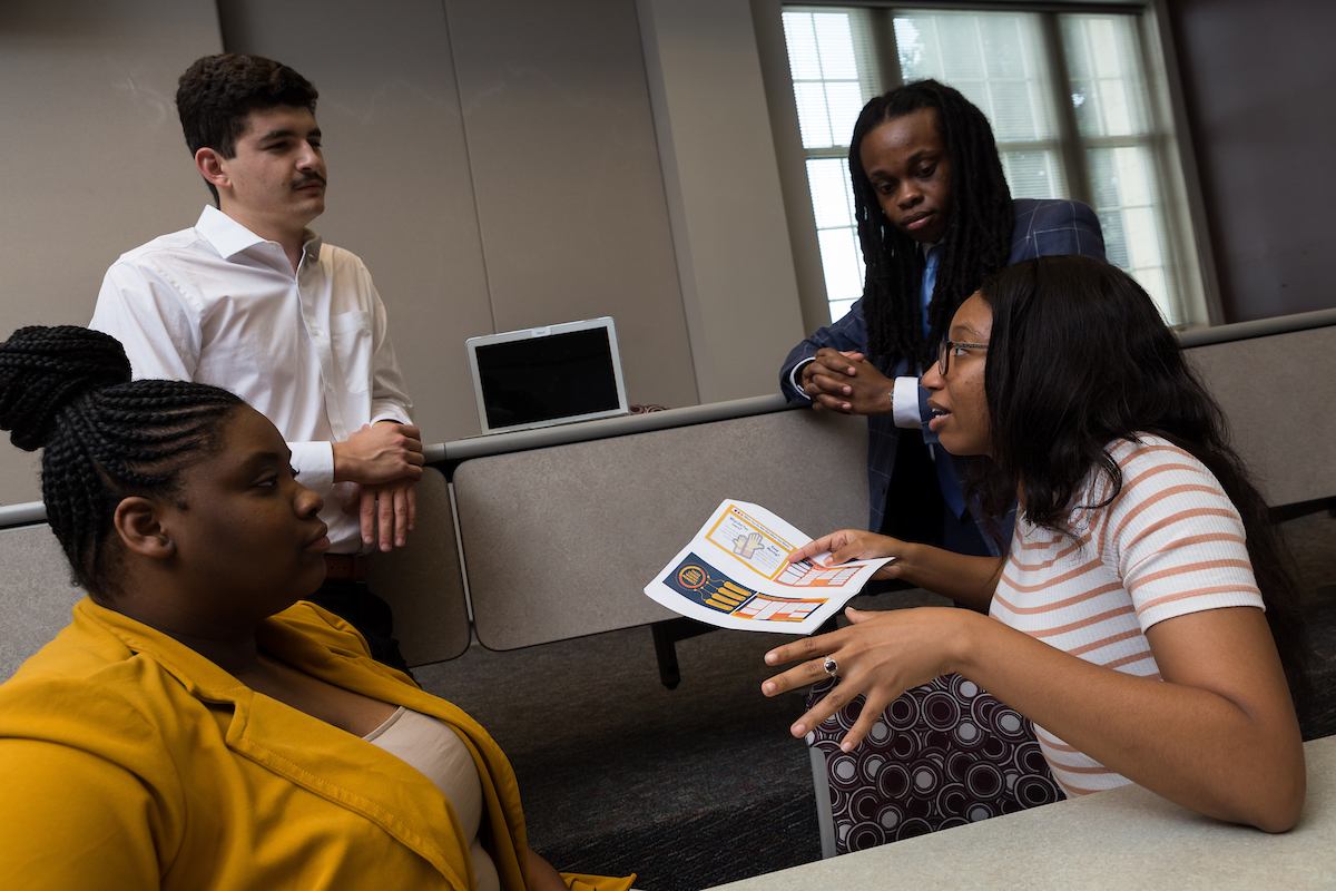 A group of four students, two seated at table, two standing, have a discussion while one holds a paper with four graphics on it
