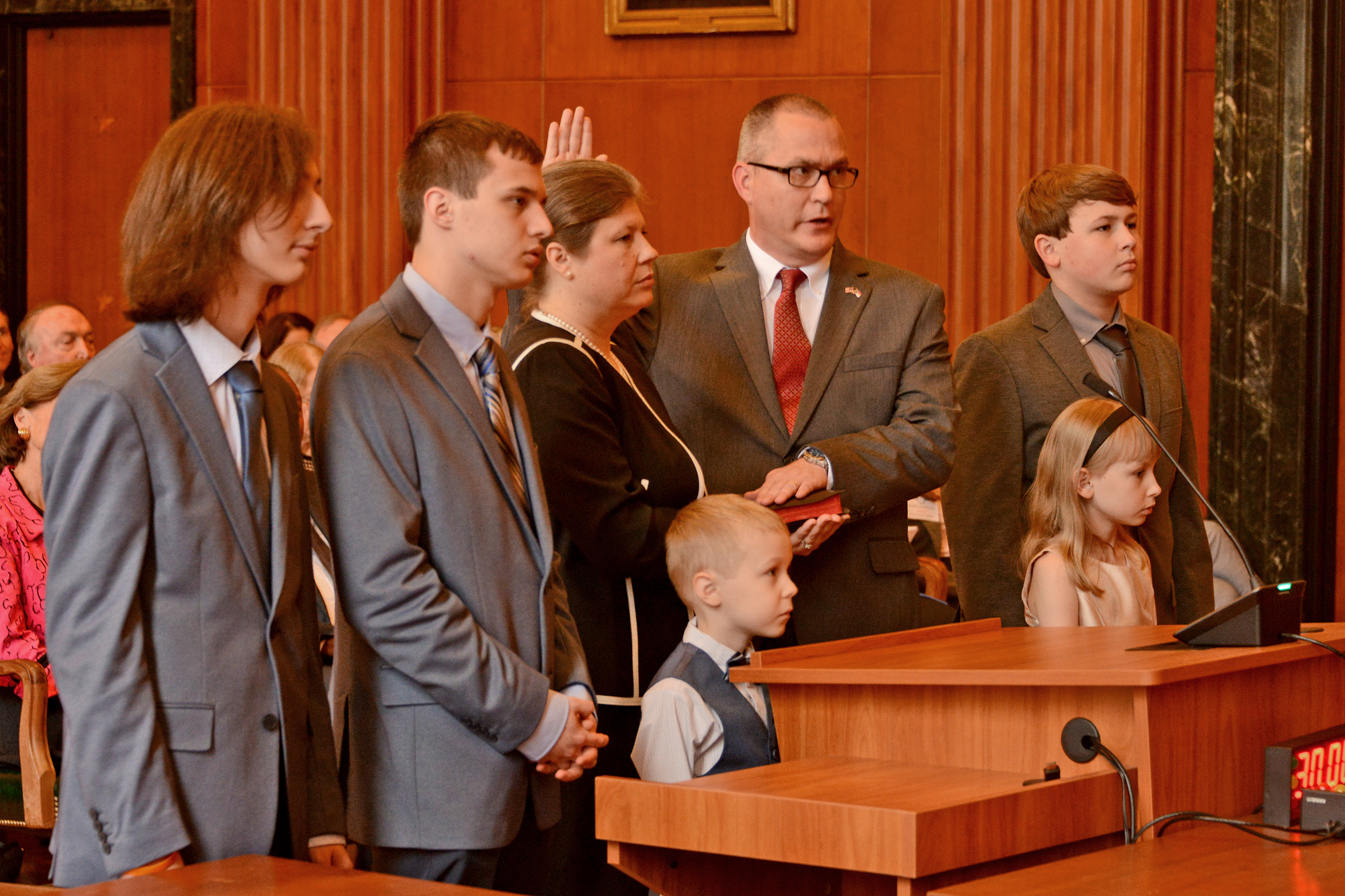 Trey Allen takes his oath of office in a formal court room with his left hand on a Bible and his right hand raised. His wife holds the Bible and they are joined by their four sons and one daughter.
