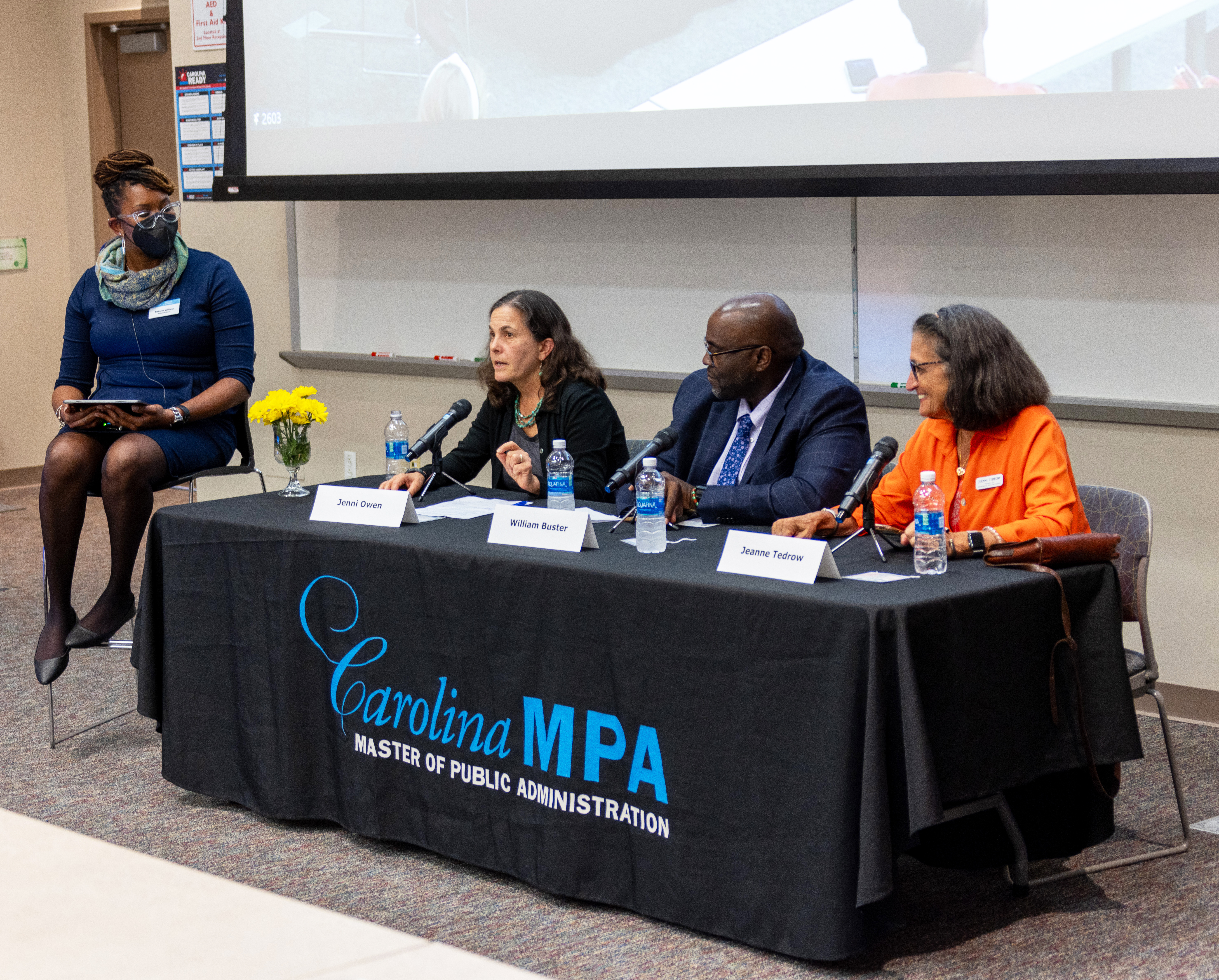 A group of 3 adults seated at a table with a banner over it that reads "Carolina MPA". One speaking into the microphone in front of her. The moderator sits in a high chair to the left of the table. 