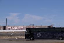 A blue tour bus drives across a concrete racetrack. A barrier in the background reads "North Wilkesboro Speedway."