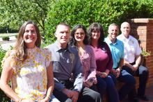 A group of six adults seated side by side on a low brick wall smiling at the camera with the sun shining on them