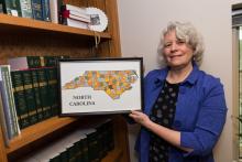 Frayda Bluestein stands in front of a bookcase filled with books. She holds a framed map of North Carolina.