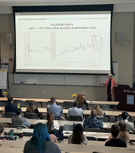 Mary Guy stands at the front of the room in front of a Powerpoint presentation. The screen reads "Voluntary Quits: Rate equals percent of total and state local government jobs" over two charts.