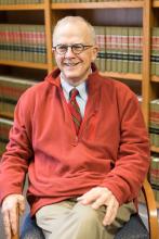 Dean Mike Smith sits in front of a bookcase of old books. He wears khaki pants and a red pullover sweater.
