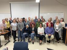 A group of 25 individuals poses for a group photo in a classroom in front of a projector screen and whiteboard.